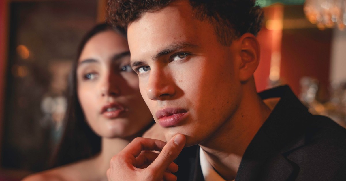 A fashionable couple in formal attire with an intense expression in a moody setting.