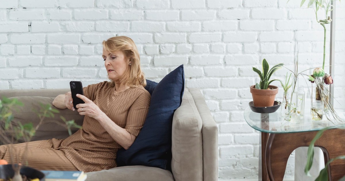 Senior female browsing internet on cellphone while resting on couch against brick wall and potted plants at home