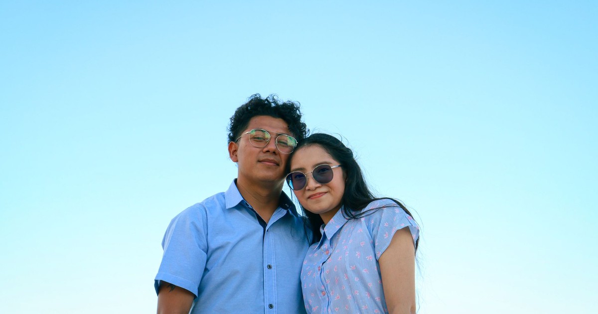 A happy couple poses together under a clear blue sky, both wearing glasses and casual attire.