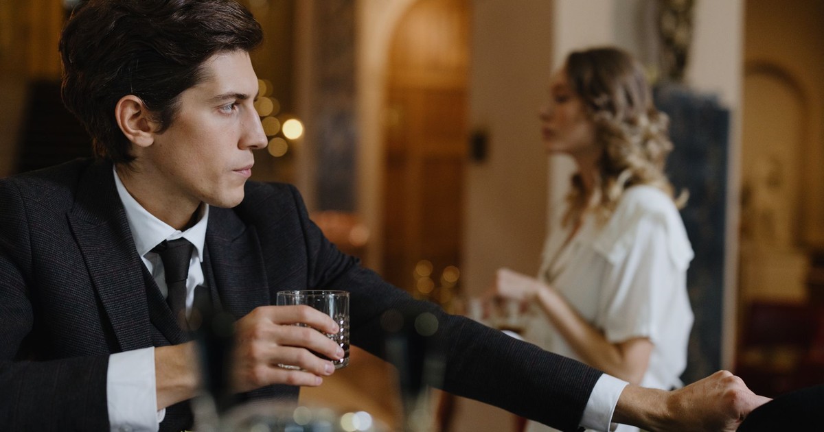 A sophisticated man in a suit holds a drink at a chic bar with a woman in the background.