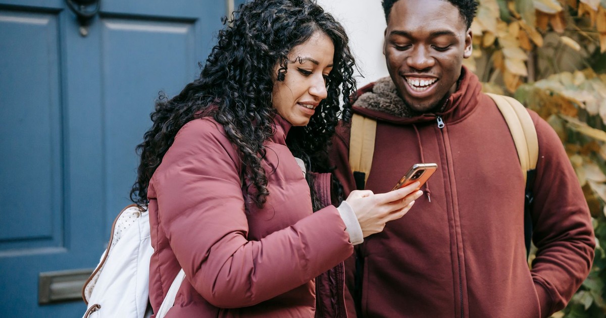 Diverse glad couple in warm outerwear using mobile phone near door of building on street