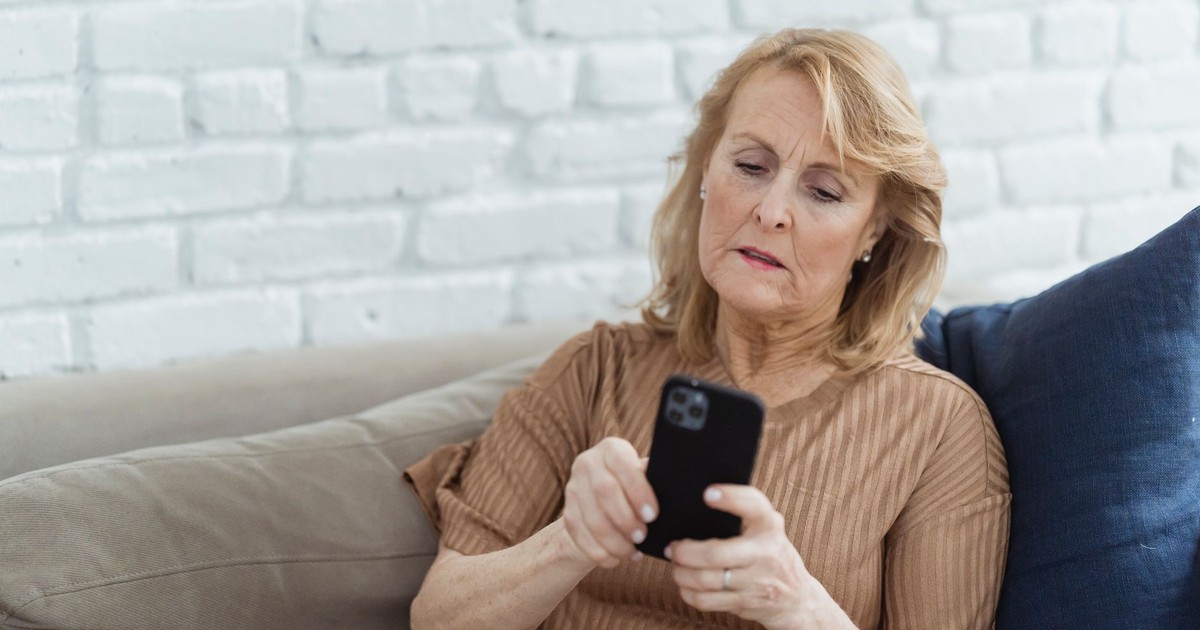 Senior female surfing internet on mobile phone while sitting on sofa against brick wall at home