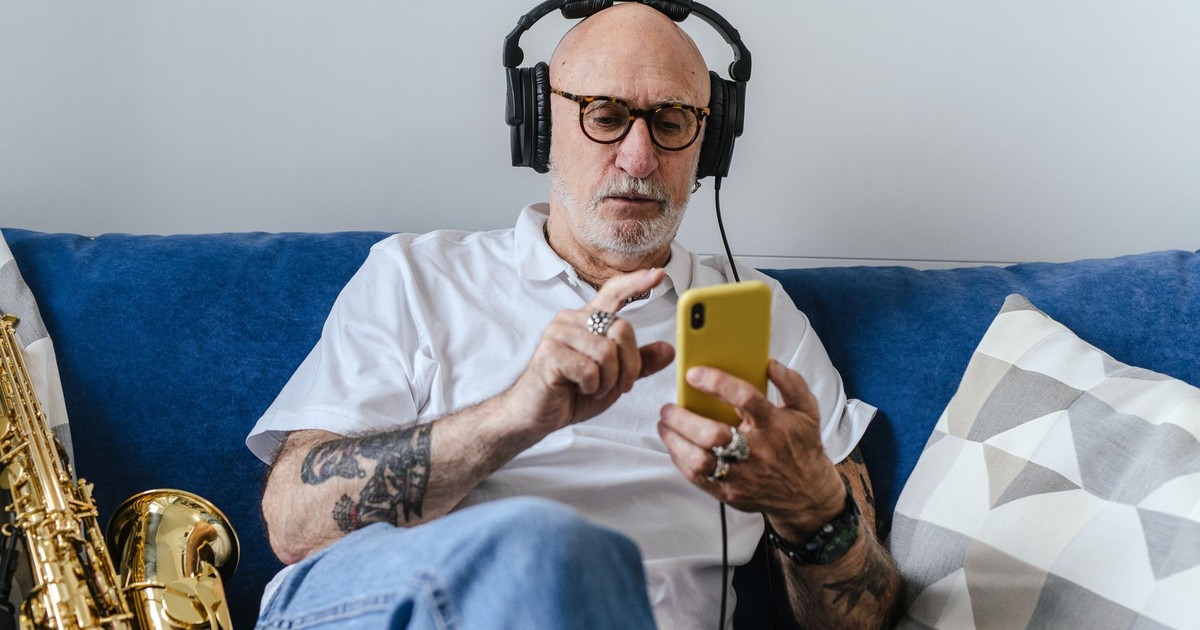 Elderly man with tattoos listening to music on headphones while using smartphone, indoor setting.