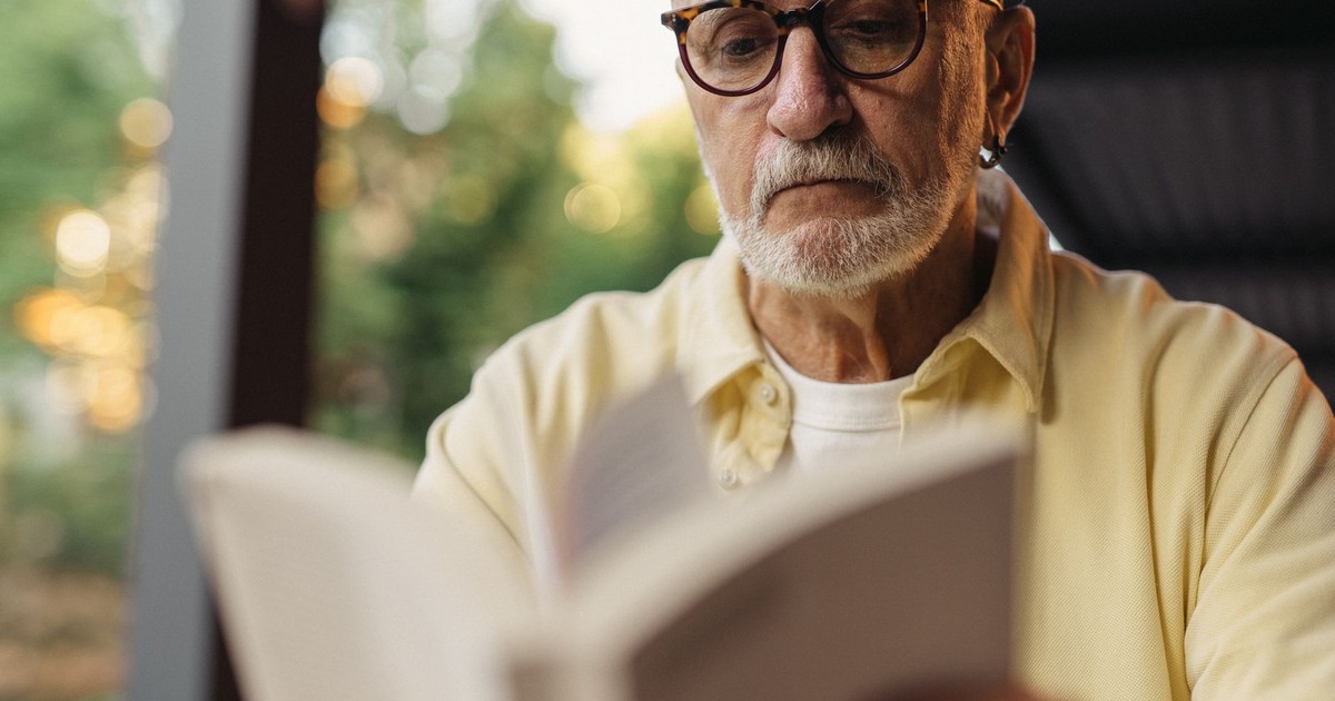 Elderly man with eyeglasses focused on reading a book outside.