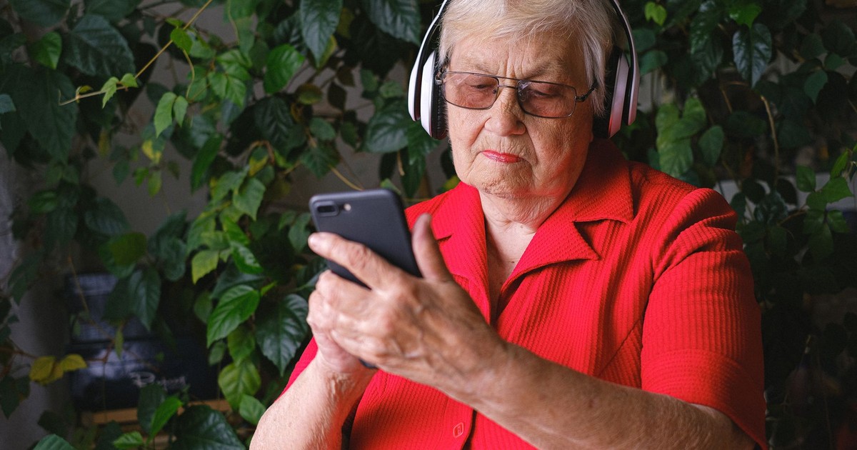 Concentrated senior female with headphones making video call online with mobile phone near fresh green leaves