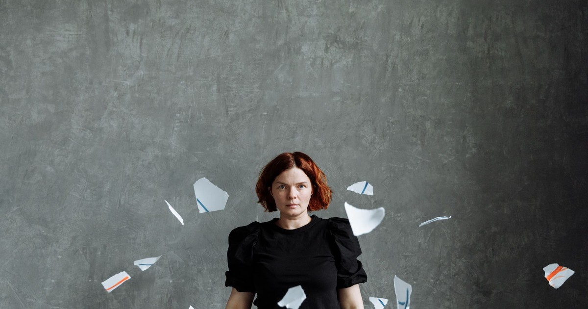 Portrait of a woman surrounded by scattered paper pieces on a gray backdrop.