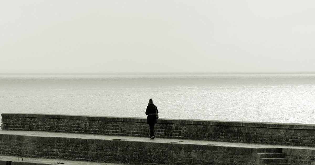 A lone figure in black and white stands by a seaside wall, evoking solitude.