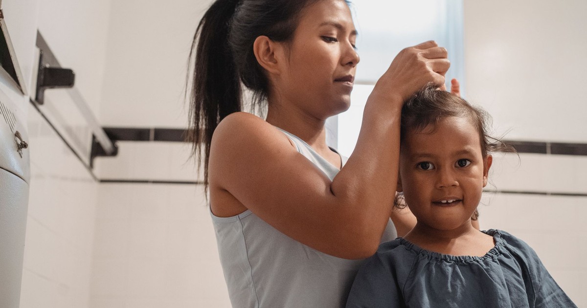 Focused young Hispanic mother in casual outfit braiding hair of cute little daughter looking at camera