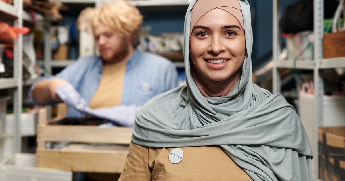 Smiling woman in hijab volunteering in a storeroom with a colleague organizing supplies.