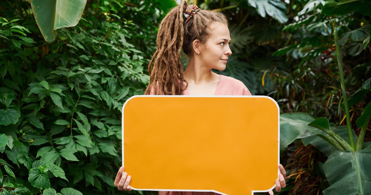 A woman with dreadlocks holds a blank orange sign in a vibrant green garden, providing perfect copy space.