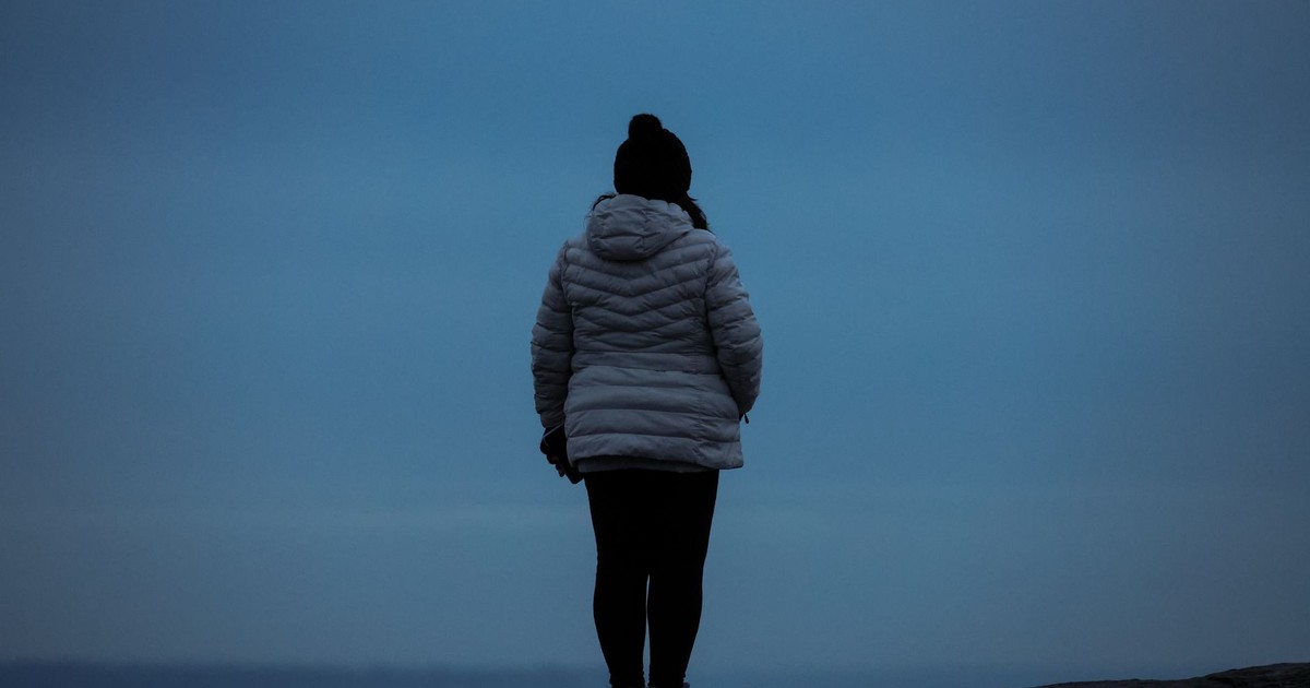 Silhouetted woman in a winter jacket standing outdoors, facing the horizon.