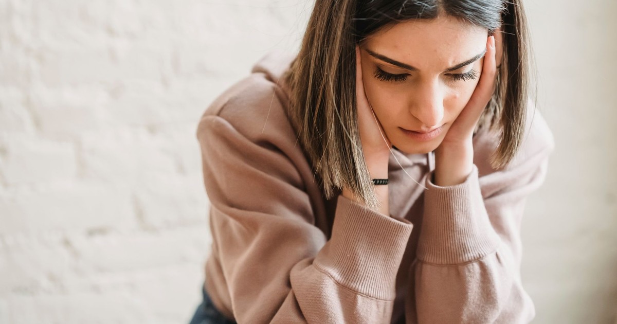 Crop unhappy female in casual wear touching cheeks and looking down while sitting near white wall in light room at home