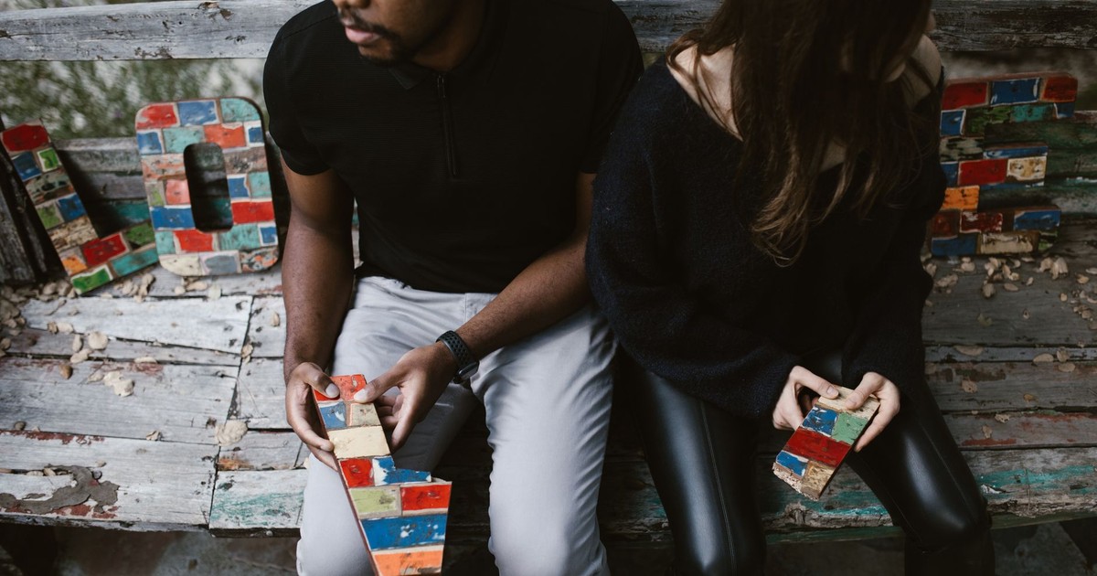 Couple sitting on a bench with colorful blocks forming the word 'LOVE,' embodying complex emotions.