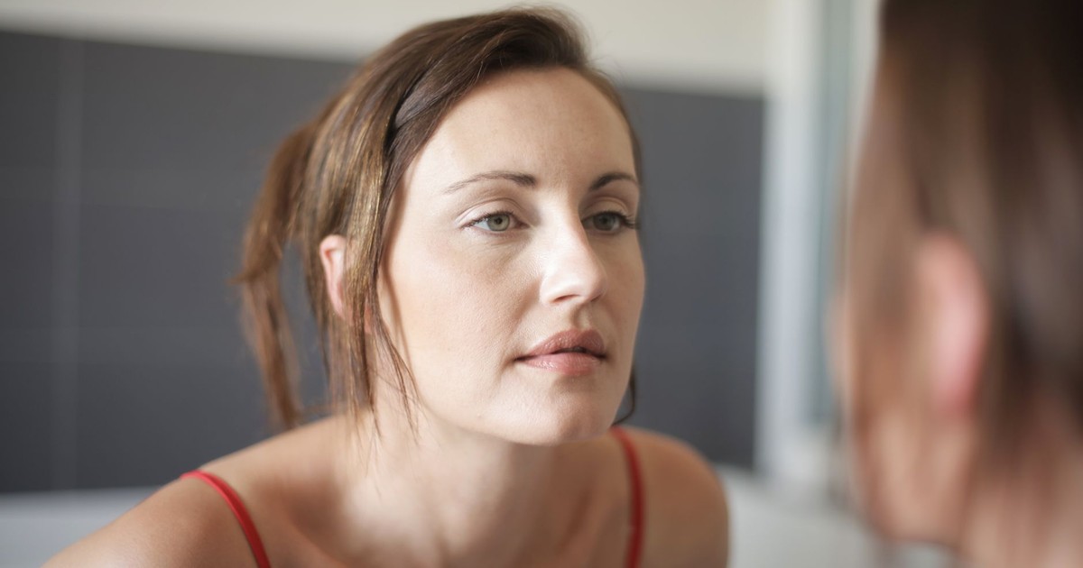 A woman thoughtfully looking at herself in a bathroom mirror.