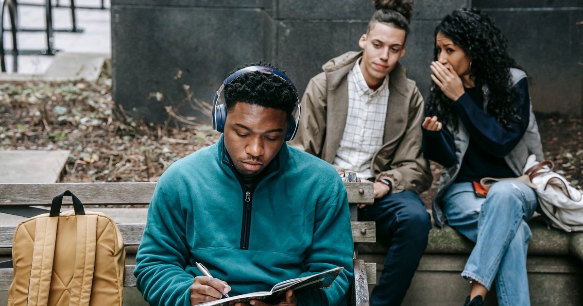 Young multiethnic friends in casual outfit with backpack gossiping behind back of African American guy sitting on bench in headphones and taking notes in notebook in city street in daylight
