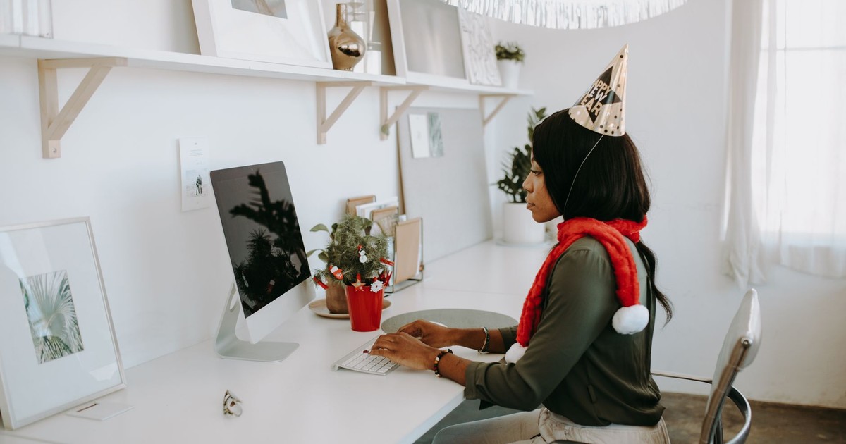 A woman in a party hat and festive scarf works at her computer in a decorated office.