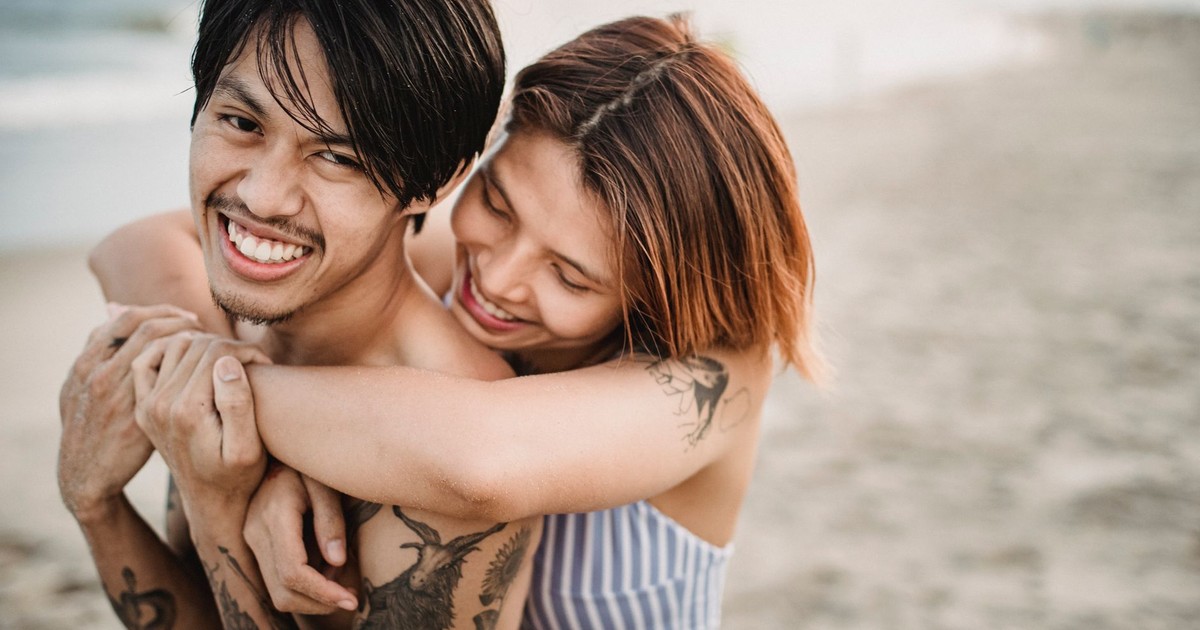 Tattooed couple smiling and hugging closely on a sandy beach during the day.