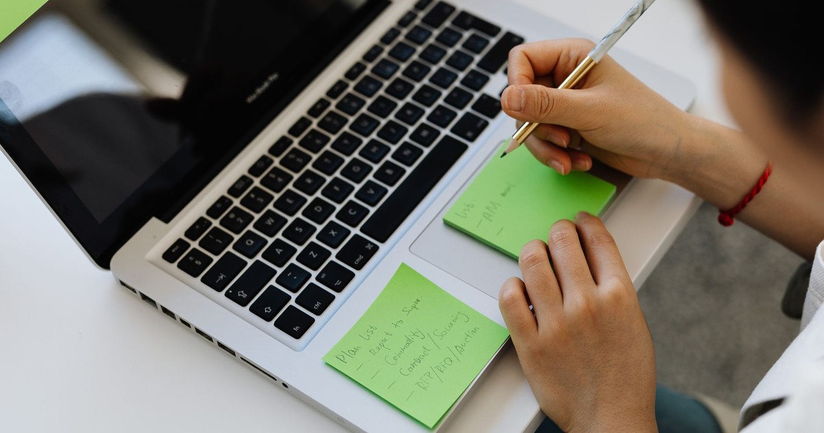 An individual writes notes on green sticky notes placed on a laptop in a work setting.
