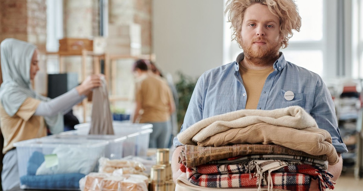 A bearded man volunteers indoors, holding donated clothes for charity work amid a collaborative environment.