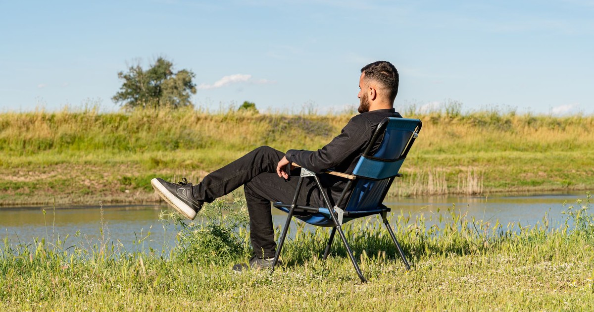 An adult man sitting leisurely in a chair by a calm riverside on a sunny day.