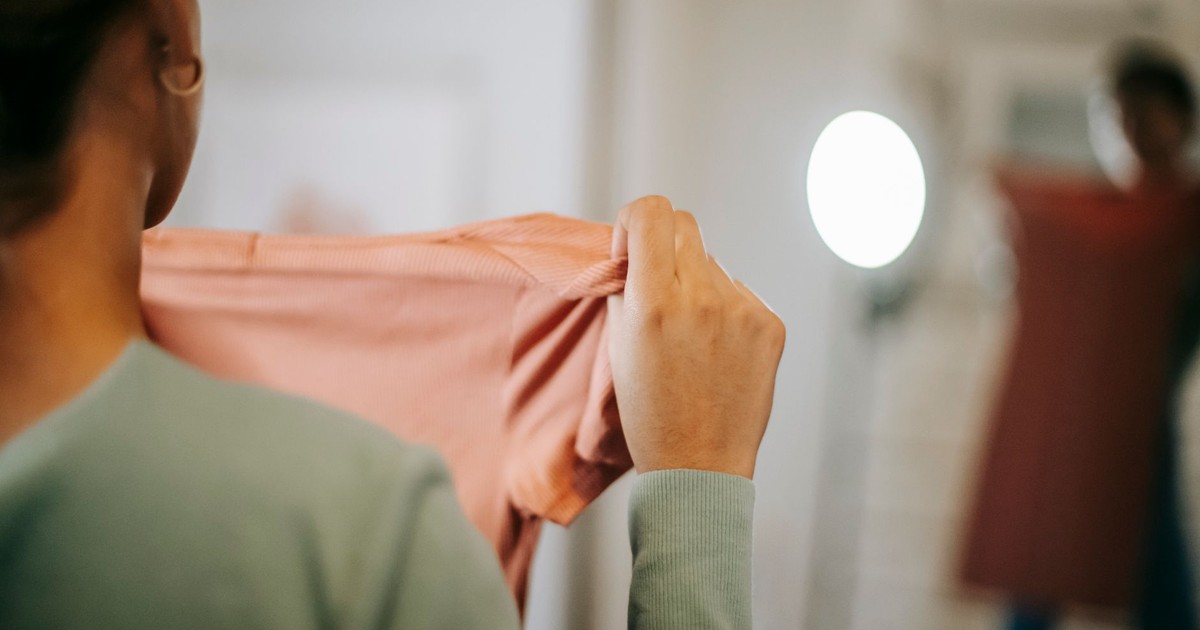 Back view of unrecognizable female in casual clothes showing pink dress while selecting clothes and looking in mirror while standing in light room at home