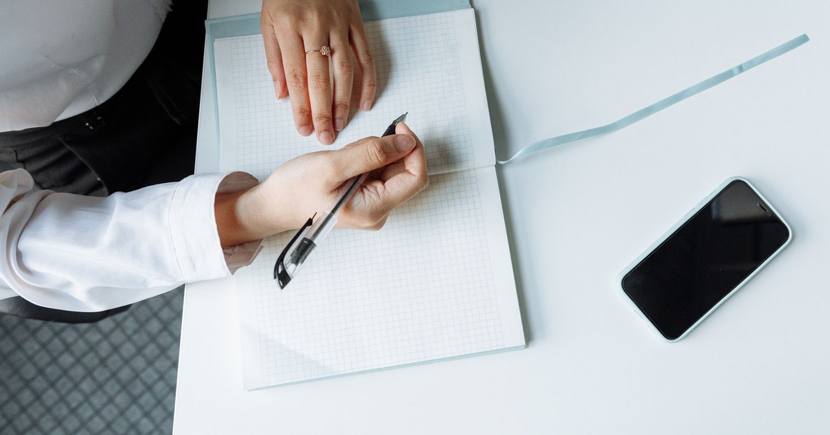 Close-up of hands writing in a notebook with a smartphone on a white desk.