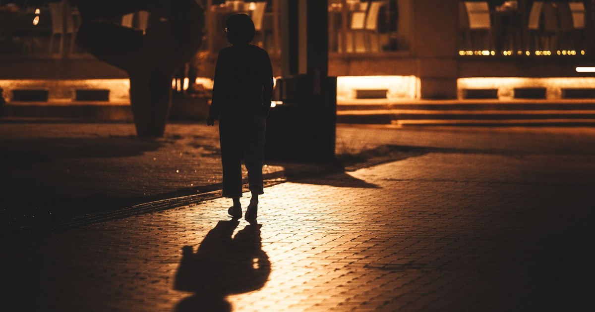 Silhouette of a woman walking on a dimly lit urban sidewalk at night, with reflections.