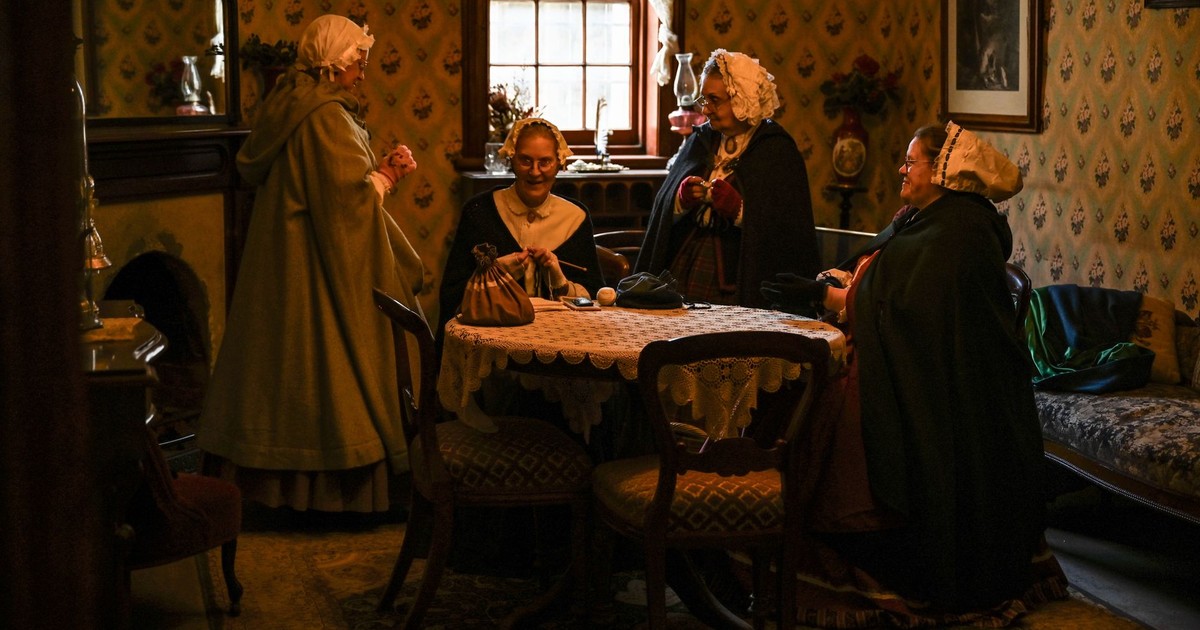 Victorian women in a cozy parlor engaged in conversation, dressed in period attire.