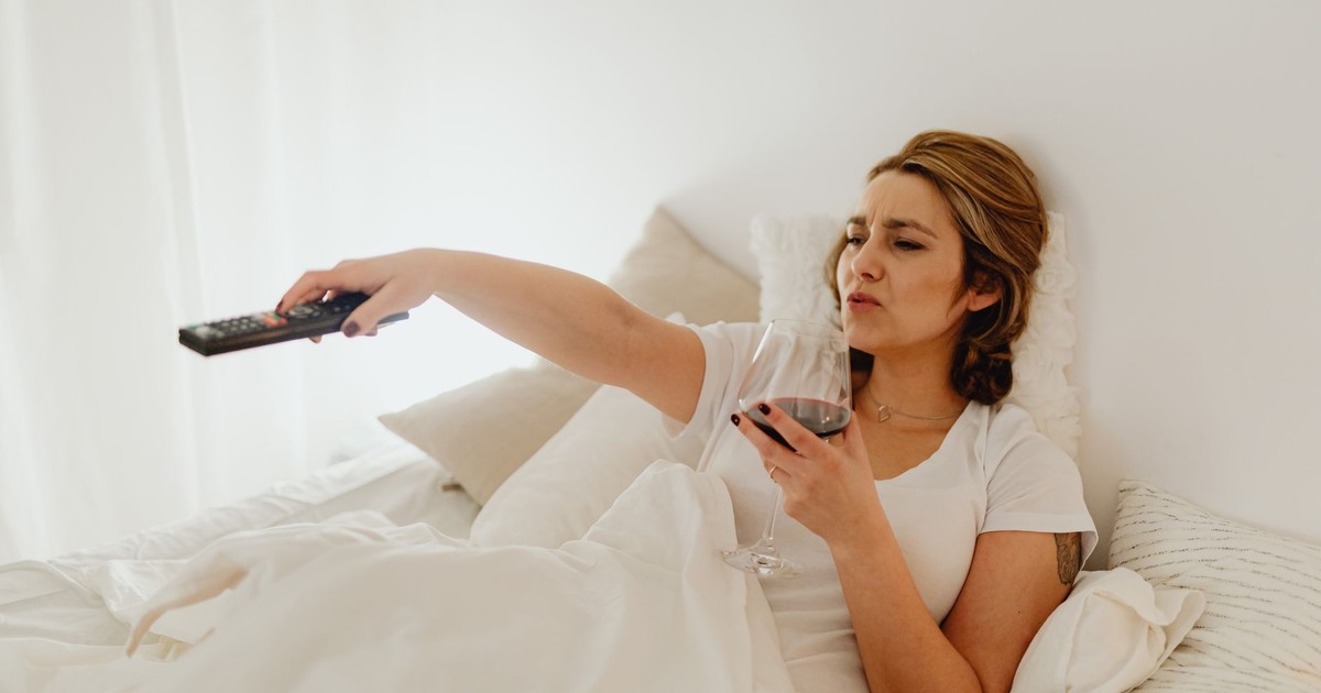 A woman in bed enjoying a glass of red wine while holding a TV remote, creating a relaxed atmosphere.
