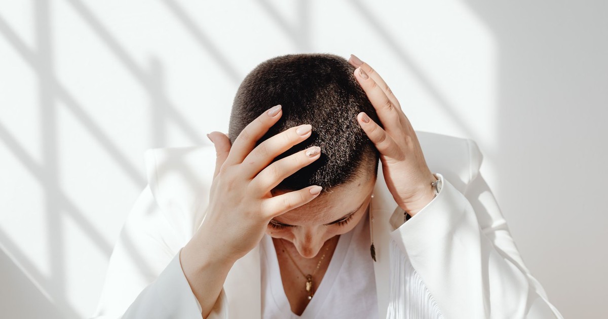Woman in white outfit feeling stressed, sitting with hands on head in bright sunlight indoors.