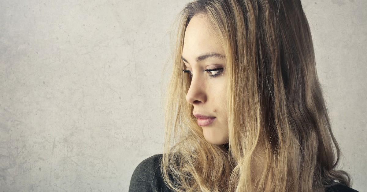 A pensive young woman with long blonde hair looking to the side against a neutral background.