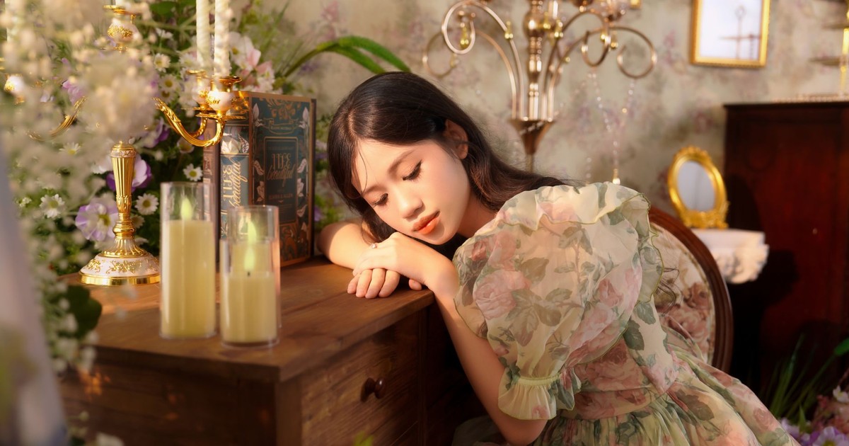 A peaceful woman in a vintage floral dress rests her head on a wooden table in an ornate room.