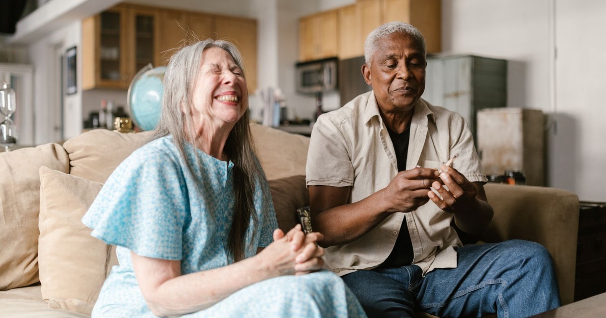 An elderly couple sitting on a couch at home, sharing a joyful moment and smiling.