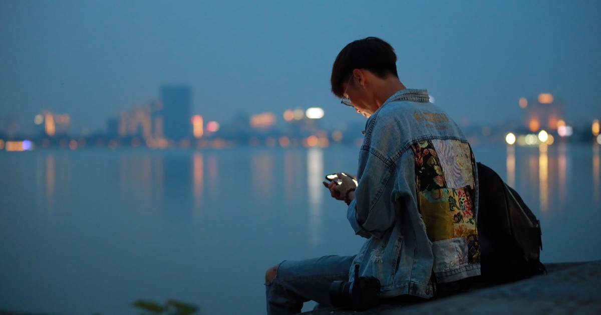 A teenager uses a phone by the waterfront at twilight, with city lights reflecting on the water.