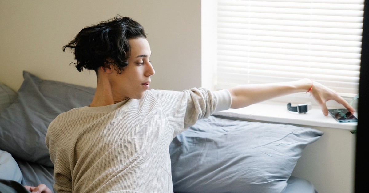 Young ethnic male adolescent taking mobile phone from windowsill after awakening in morning in bedroom
