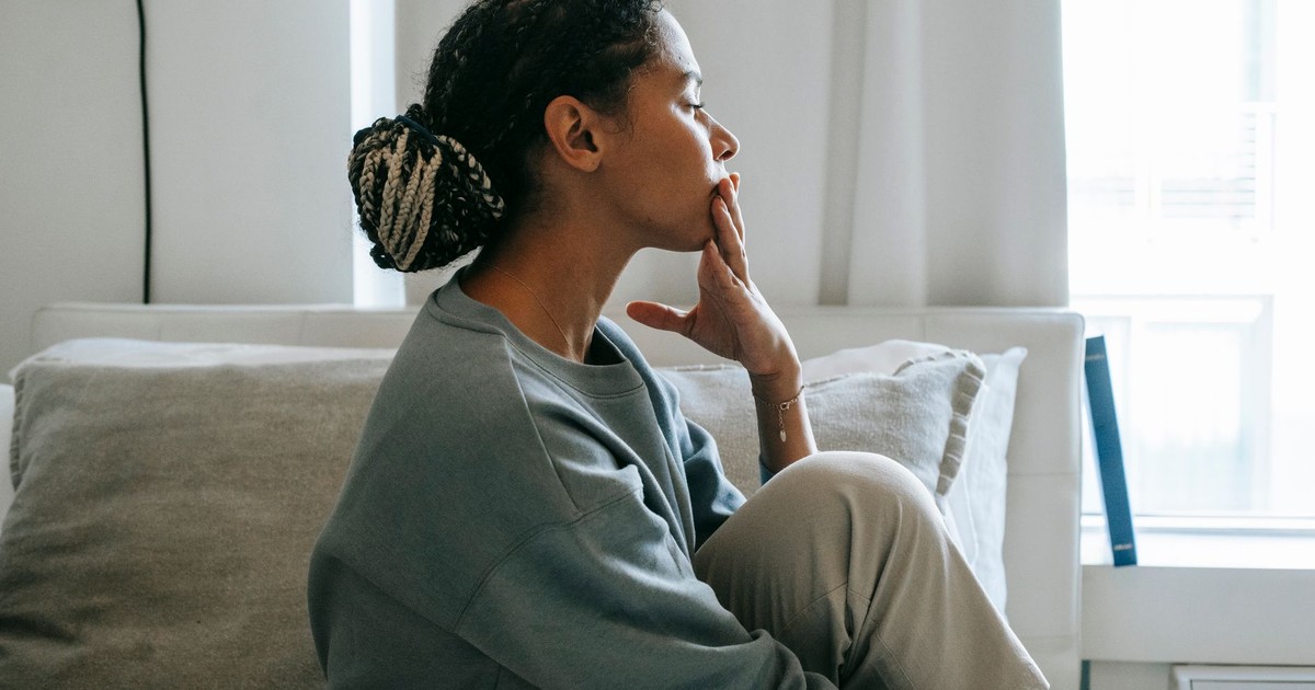Side view young African American woman sitting on comfortable white bed and thinking in casual clothes in daytime