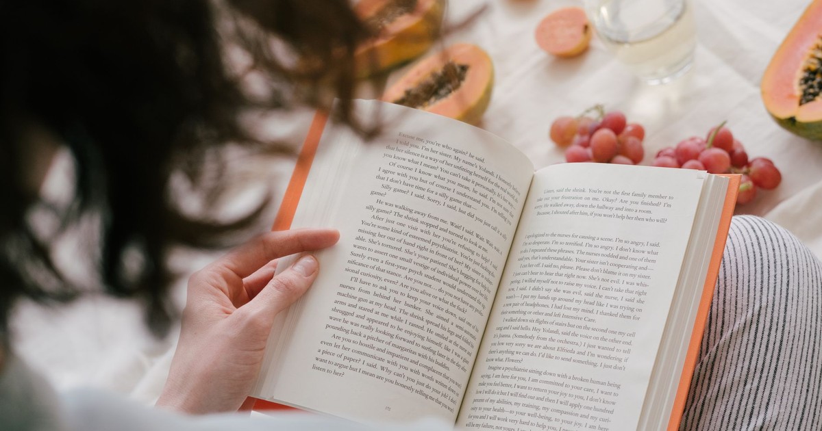A person enjoys reading a book surrounded by fresh papaya, grapes, and a drink, creating a cozy indoor scene.