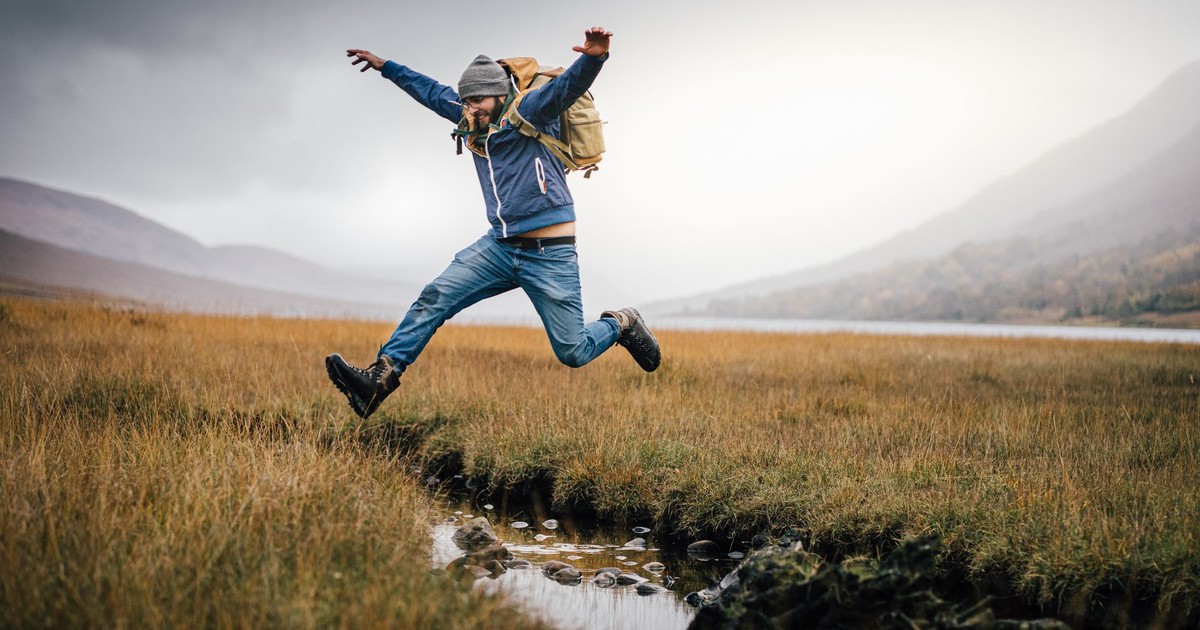 A man jumps with excitement over a stream in a picturesque, open landscape.