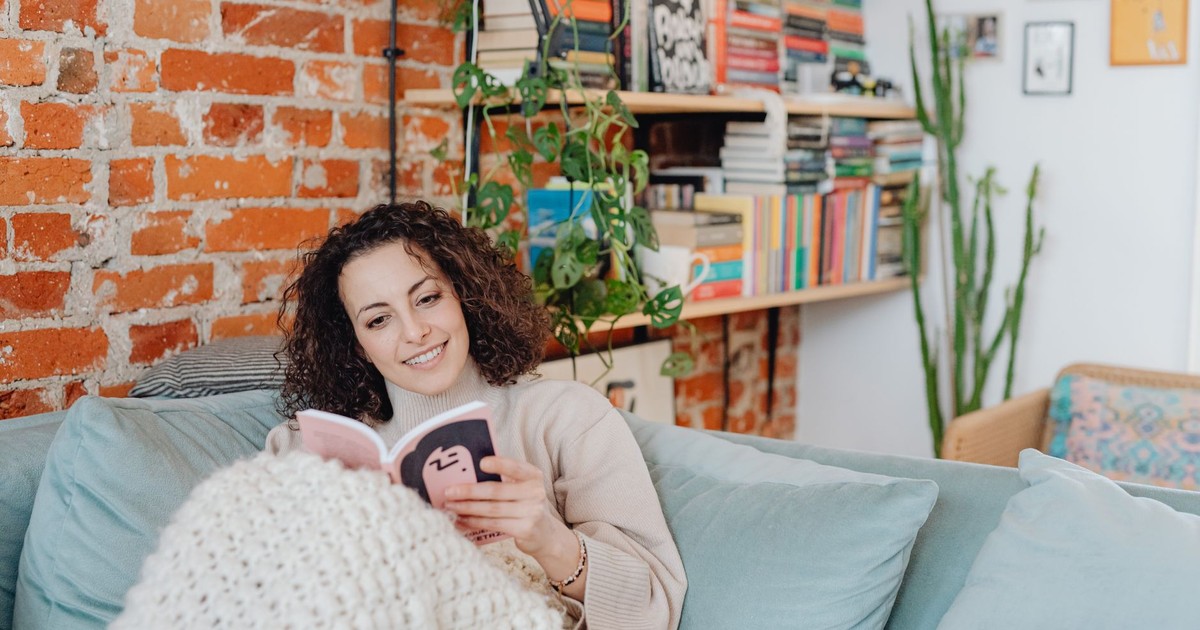 A smiling woman reads on a sofa in a cozy room with bookshelves and plants.
