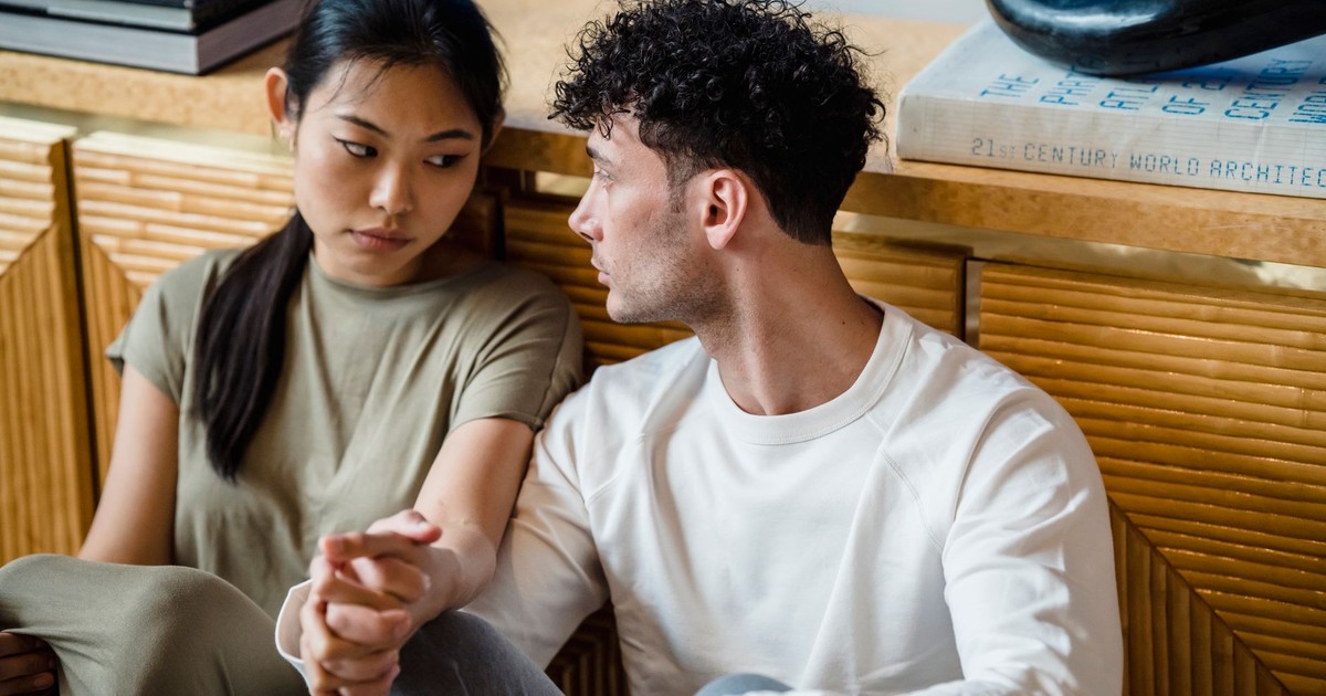 A couple sitting together indoors, holding hands with thoughtful expressions.