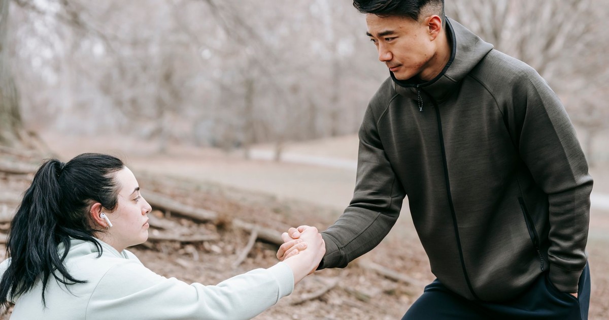 Young Asian male in sportswear giving hand to plump female fell on ground while training together in autumn leafless forest