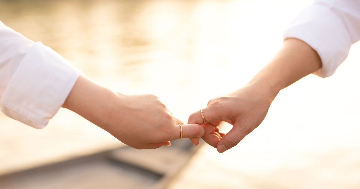A tender moment between a couple touching hands by the water, showcasing wedding rings in soft sunlight.