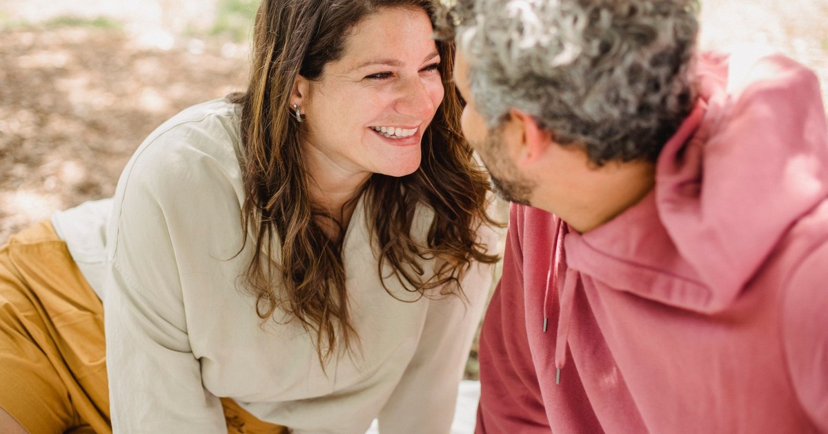Smiling pregnant female with male sitting on grass and looking at each other on sunny summer day