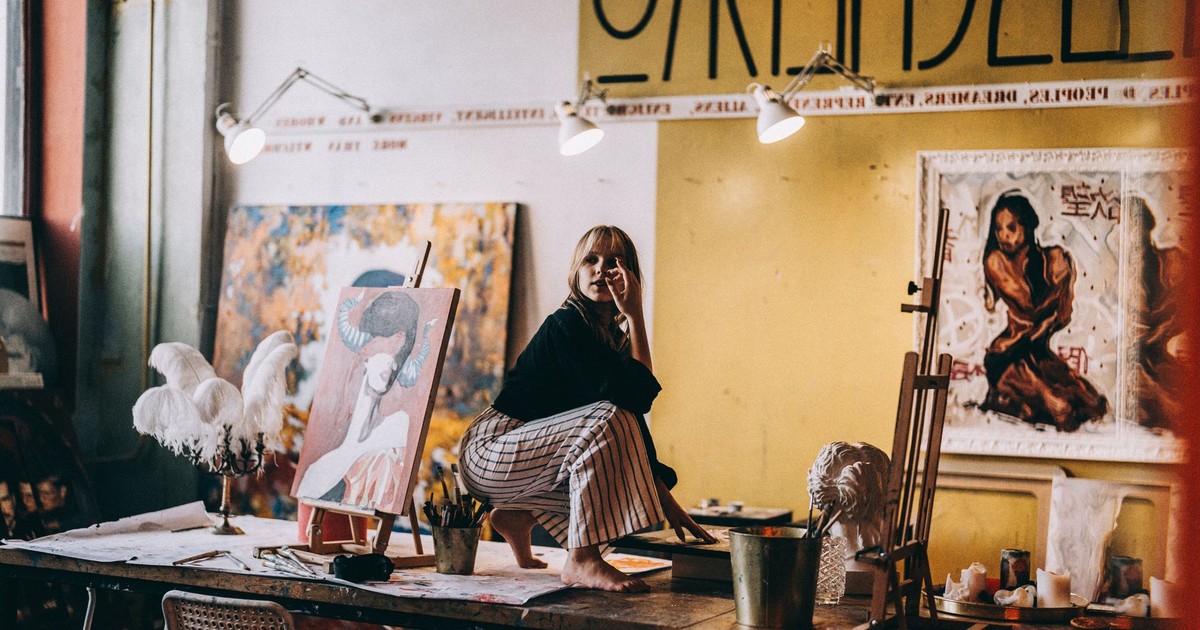 A woman artist sitting on a work table in a vibrant art studio surrounded by paintings and sculptures.