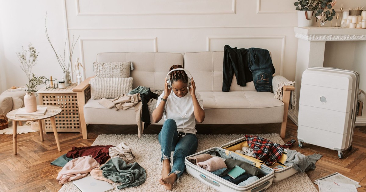 A woman packs a suitcase in her living room while listening to music, surrounded by clothes and a cozy atmosphere.