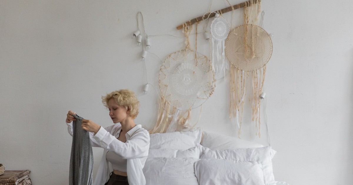 Tranquil female with blond hair sitting on bed and putting garment in suitcase while preparing for travel in cozy room