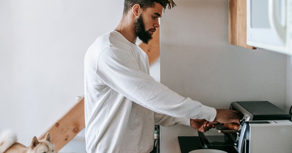 Side view of stylish young African American male owner in casual clothes heating croissant in microwave oven while standing in kitchen with cute Akita Inu dog