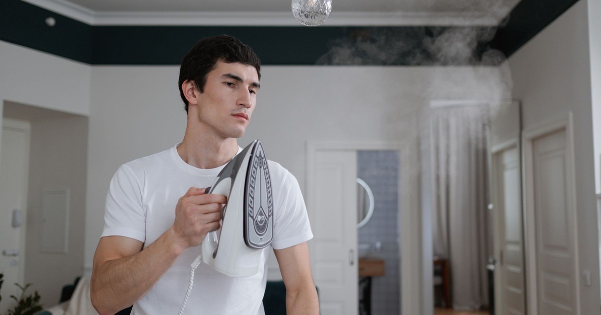 A man uses a steam iron on a white shirt indoors, highlighting daily household chores.