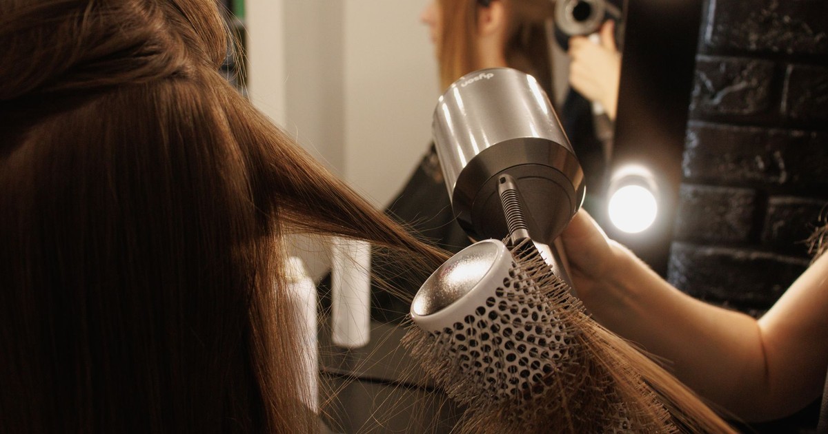 A stylist blow-dries a woman's hair using a brush and hairdryer in a salon.