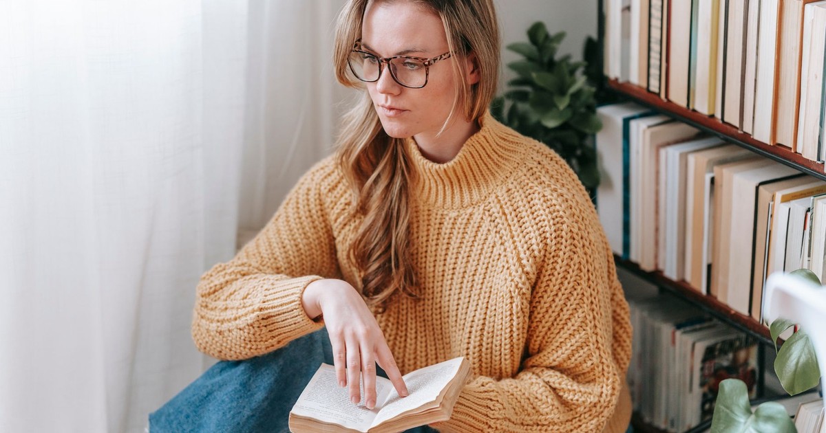 Pensive female student in casual clothes and eyeglasses sitting near bookshelf while reading book in light apartment